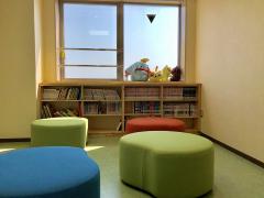 図書コーナー写真。床はモスグリーン、壁はクリーム色。窓からの採光あり部屋は明るい。子どもならば何人も座れるような低い背もたれのないカラフルなソファが4つ写っている。備え付けの気の棚には子供向けの本がたくさん準備されている。棚の上にはぬいぐるみが数体おかれて、穏やかな雰囲気をかもし出している。