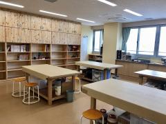 クラフトルーム写真。壁まである備え付けの木製の棚。上の2段は扉がついている。4人がけの作業台には丸椅子がある。