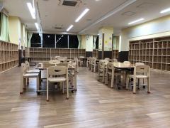 学童保育クラブの室内写真。床と棚は木材。ランドセルなどが入る大きな棚。6人がけのテーブルと椅子が設置されていて、工作などで使うであろう電源を取るためのケーブルが天井からぶら下がっている。