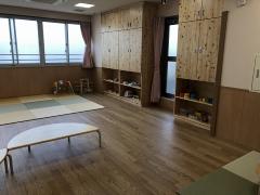 乳幼児遊戯室の写真。大きな窓から採光して部屋は明るい。床はフローリング。備え付けの棚も木目でやさしい印象。四角い組み合わせることの出来るマットも用意され、寝転がってもすごせるようになっている。乳幼児向
