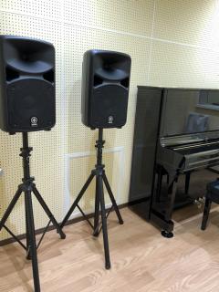 スタジオの写真。左手前スタンド型スピーカー2台と右奥にピアノがある。